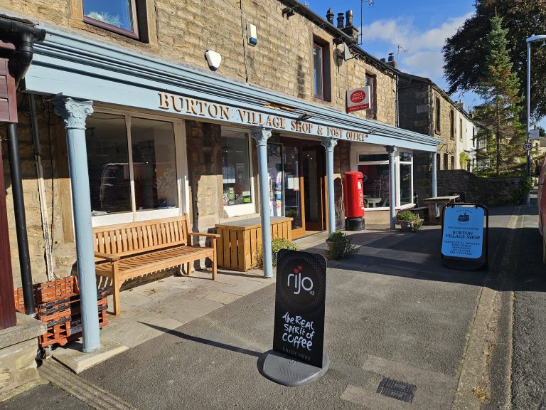 View of Burton village shop. The shop is on the right hand side of the picture, with the camera looking down the pavement towards houses behind it.